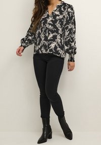 Blouse noire à motifs floraux avec manches longues, poignets élastiques, col en V et motif de feuilles crème, associée à un jean skinny noir et des bottines.