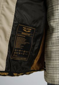 Doudoune avec un tissu beige et noir, comportant une étiquette dorée avec des spécifications. Intérieur doublé d'un matériau jaune et avec des accents texturés.