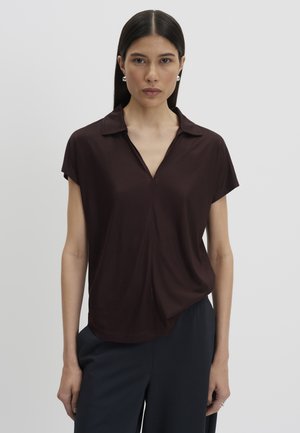Donna con capelli lisci e scuri indossa una maglia marrone scuro a maniche corte con colletto e pantaloni scuri, in piedi contro uno sfondo grigio chiaro.
