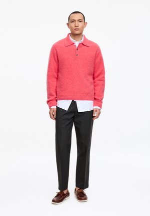 Hombre con suéter rosa coral sobre camisa blanca, pantalones negros y zapatos náuticos de cuero marrón, de pie sobre fondo blanco liso.