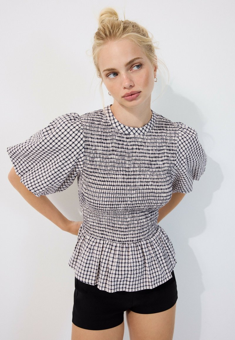 Femme aux cheveux blonds en chignon, portant un haut à manches bouffantes à carreaux noir et blanc et un short noir, debout les mains dans le dos.