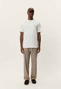 Λευκό κοντομάνικο μπλουζάκι με στρογγυλή λαιμόκοψη και μικρό σχέδιο κειμένου στο στήθος, συνδυασμένο με ανοιχτό καφέ καρό παντελόνια και σκούρα παπούτσια.