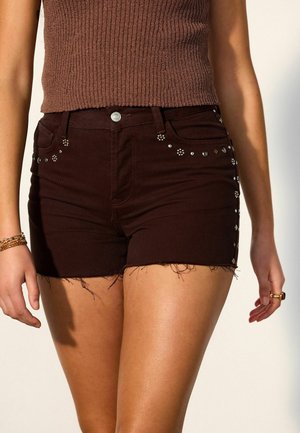 Woman wearing dark brown studded denim shorts with frayed hems and a sleeveless ribbed brown top, standing with arms relaxed.