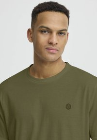 T-shirt verde oliva a maniche corte realizzata in tessuto liscio, con un piccolo logo geometrico nero sul petto e un collo rotondo.