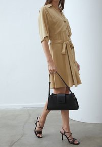Beige shirt jurk met een riem, korte mouwen en twee voorkruiden, gecombineerd met een zwarte handtas en zwarte sandalen met bandjes. Glad textiel.