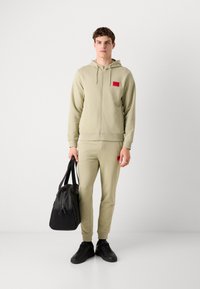 Light green zip-up hoodie with red logo, matching pants, and black sneakers; holding a black duffel bag. Smooth fabric, casual fit.