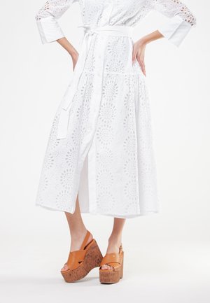 Donna che indossa un vestito midi bianco con motivo ad occhiello, cintura annodata in vita e sandali zeppa marroni in pelle con suole in sughero.