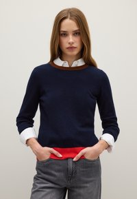 Maglione a maglia blu navy con collo a coste marrone e orlo rosso, indossato sopra una camicia bianca con colletto, abbinato a jeans grigi.