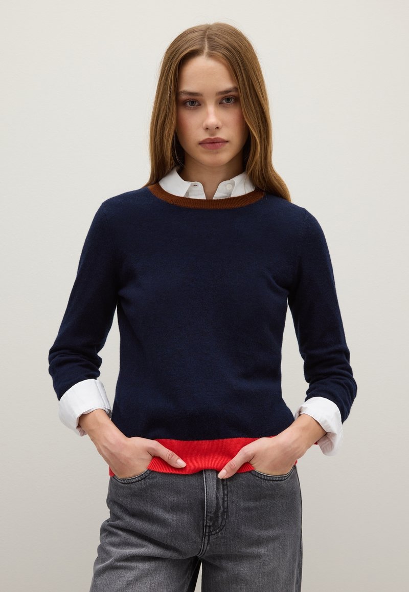 Maglione a maglia blu navy con collo a coste marrone e orlo rosso, indossato sopra una camicia bianca con colletto, abbinato a jeans grigi.