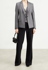Blazer gris sur mesure avec un col cranté porté sur un gilet assorti, accompagné d'un pantalon noir large. Un sac à main noir à la main complète la tenue.