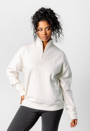 Frau mit lockigem Haar, die einen weißen Sweatshirt mit Reißverschluss bis zur Brust und dunkle Hosen trägt, steht mit einer Hand in der Hüfte und schaut seitlich.