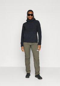 Zwarte fleece-top met hoge kraag, groene outdoor-broek met een logo, en zwarte schoenen. Glad textuur, getailleerd silhouet, functioneel ontwerp.