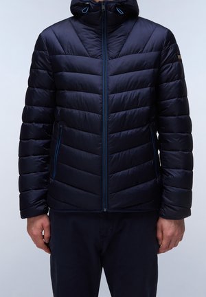 Uomo che indossa una giacca imbottita blu navy con cappuccio e tasche con cerniera, in piedi davanti a uno sfondo semplice.