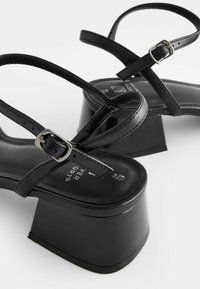 Sandales noires à talons carrés épais, bouts carrés et fines brides ajustables à la cheville avec boucles argentées, présentées sur un fond blanc.