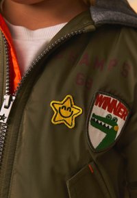Veste bomber vert olive avec une capuche grise, un patch étoile jaune avec un visage souriant, et un patch brodé "WINNER" avec un motif de crocodile.
