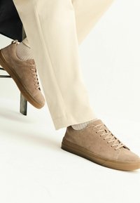 Baskets en daim marron clair avec lacets plats et semelles en caoutchouc. Portées avec un pantalon beige et des chaussettes gris clair, mettant en avant un design minimaliste.