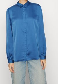 Blå satin långärmad skjorta med spetsig krage, knapp-up-front och volangmanchetter, tillsammans med ljusblå jeans.