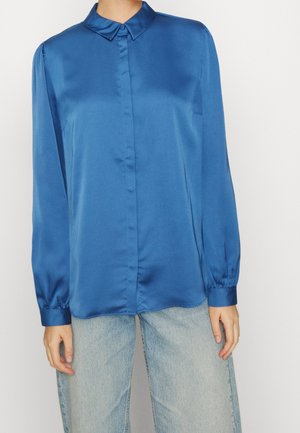 Camicia lunga in satin blu con colletto punta, chiusura con bottoni e polsini arricciati, abbinata a jeans chiari.