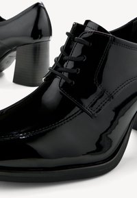 Scarpe con tacco in vernice nera, con punta affusolata, design con lacci e tacco in gomma nera testurizzata. Finitura liscia, dettagli di cucitura minimi.