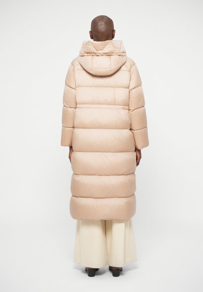 Hosszú bézs színű puffertakaró kapucnival, vízszintes steppelt részekkel, széles ujjakra és sima, fényes felületre jellemző.