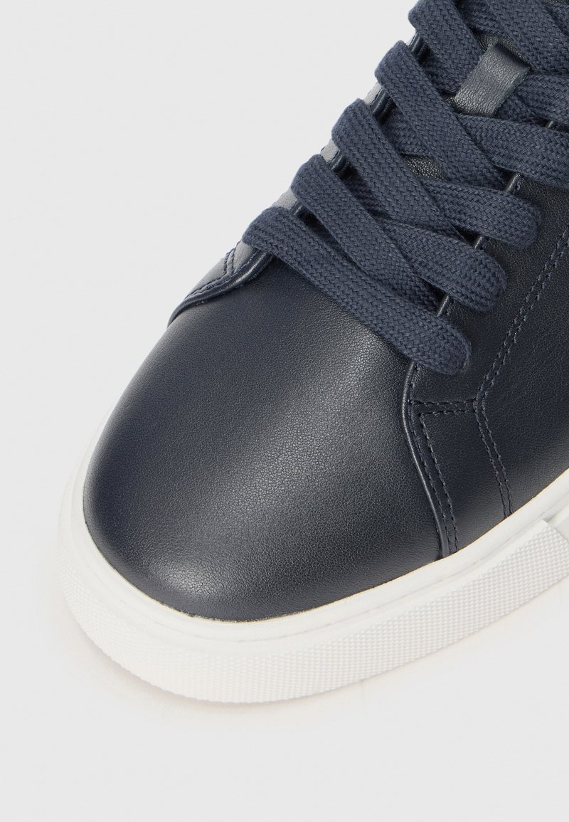 Acercamiento de una zapatilla de cuero azul marino con cordones azul oscuro y suela de goma blanca sobre un fondo claro.