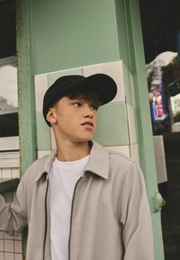 Grå zip-up jacka, vit t-shirt under, svart keps, står mot en grön och vit kaklad vägg med synliga rostiga detaljer.