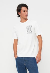 T-shirt en coton blanc avec un imprimé graphique noir sur la poitrine gauche. Manches courtes et col rond, associé à un jean en denim bleu.