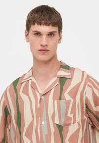 Camisa de botones de manga corta en tejido ligero, con un patrón de zigzag en rosa, verde y beige, y un bolsillo en el pecho.
