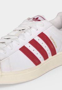 Baskets blanches Adidas Superstar avec des bandes rouges, texte doré « SUPERSTAR », lacets blancs et semelle crème sur un fond clair.