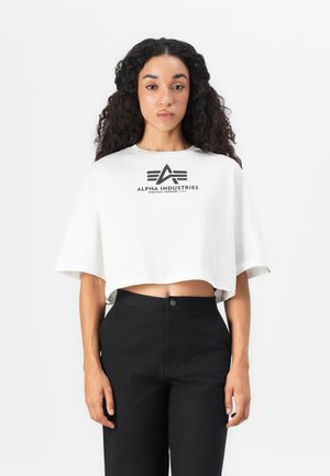 Jeune femme avec de longs cheveux bouclés, portant un t-shirt cropped blanc d'Alpha Industries et un pantalon noir taille haute, se tenant devant un fond blanc.