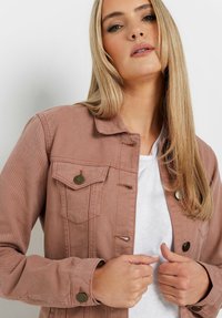 Veste en denim rose clair dotée d'un devant à boutons, de deux poches poitrine et de boutons en métal ronds. Portée par-dessus un haut blanc. Texture lisse.