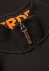 Felpa nera con zip e colletto strutturato, con cerniera argentata e un tirazip in pelle. La parola "SUPERDRY" è ben visibile in arancione.