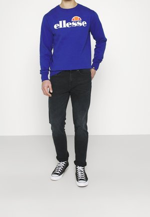 Sweatshirt bleu à manches longues avec le logo "ellesse", associé à un jean skinny noir et des baskets noires avec des semelles blanches. Fond uni.