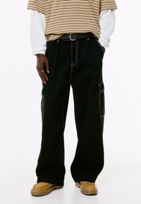 Personne portant un pantalon cargo noir à jambes larges avec des coutures blanches, des bottes beiges, une chemise rayée beige sur une chemise à manches longues blanche, les mains dans les poches.