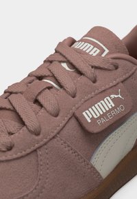 Κοντινό πλάνο της σκόνινης ροζ αθλητικής παπουτσας Puma Palermo με υφασμένα κορδόνια, λευκή ετικέτα με το λογότυπο και λευκή λωρίδα στο πλάι πάνω σε σόλα από καουτσούκ.