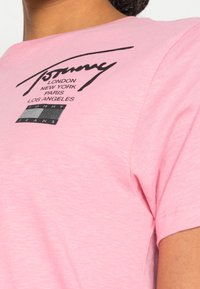 Růžové bavlněné tričko s kulatým výstřihem, krátkými rukávy a černým grafickým logem se jmény měst a nápisem "TOMMY JEANS" na přední straně.