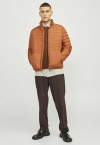 Chaqueta acolchada marrón con un diseño acolchado, llevada sobre un suéter marrón y una camisa de cuadros, combinada con pantalones oscuros y zapatos negros.