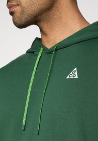 Πράσινο φούτερ με υφή, που διαθέτει τριγωνικό λευκό λογότυπο και αντίθετα πράσινα κορδόνια. Κοντινή λήψη της περιοχής του λαιμού.