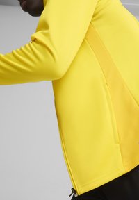 Gelbe Sportjacke mit langen Ärmeln, verfügt über ein strukturiertes Rückenpanel, eine seitliche Reißverschlusstasche und einen glatten Stoff mit enganliegendem Design.