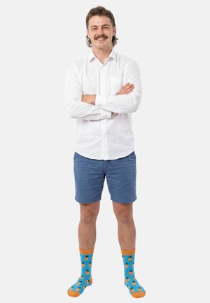 Man with mustache standing with arms crossed, wearing white button-up shirt, blue shorts, and blue socks with orange pineapples.