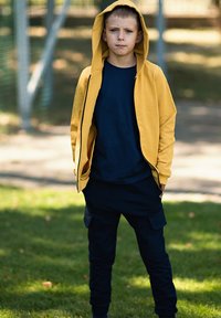 Żółta kurtka z kapturem na ciemnoniebieską koszulę, zestawiona z ciemnoniebieskimi spodniami bojówkami. Tkanina jest gładka, o luźnym kroju z bocznymi kieszeniami.