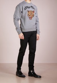 Felpa grigia con grafica di leopard e testo "Stay No", abbinata a pantaloni neri aderenti e stivaletti neri, in un contesto minimalista.