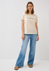 Beige t-shirt med kort ärm med trycket "Vandra och var nyfiken", kombinerad med vida ljusblå denimjeans och bruna sandaler.