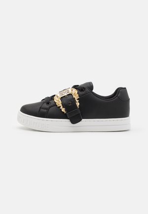 Baskets en cuir noir avec semelle extérieure blanche, dotées d'une boucle décorative en or marquée "Versace Jeans Couture". Texture lisse et design à bout rond.