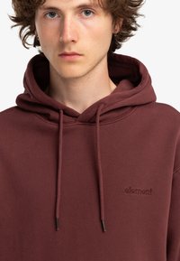 Bordeaux hættetrøje lavet af blødt stof, med en frontkangaroo-lomme, snorhætte og subtilt broderet logo på brystet.