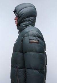 Giubbotto puffer di colore verde scuro, caratterizzato da un design trapuntato, un ampio cappuccio e una finitura testurizzata. La manica sinistra presenta una patch del marchio.