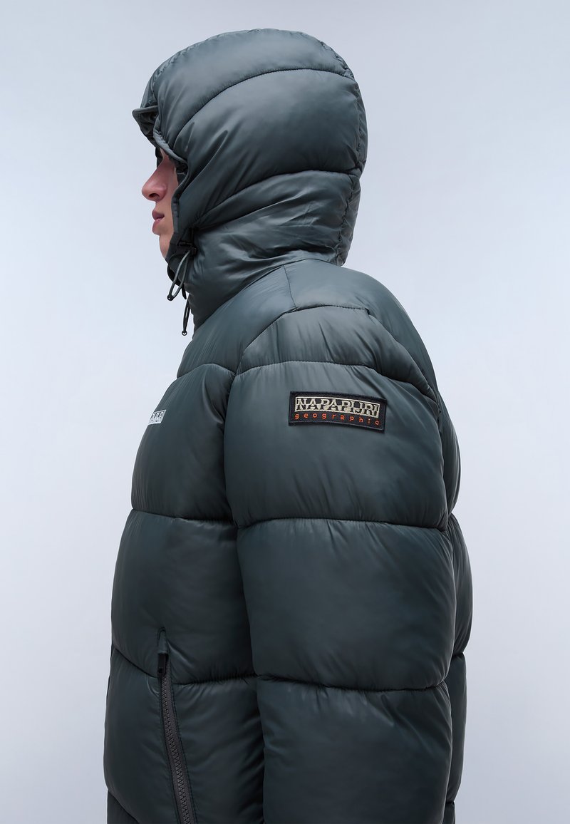 Dunkelgrüne Pufferjacke mit einem gesteppten Design, einer großen Kapuze und einer strukturierten Oberfläche. Der linke Ärmel zeigt ein Markenpatch.