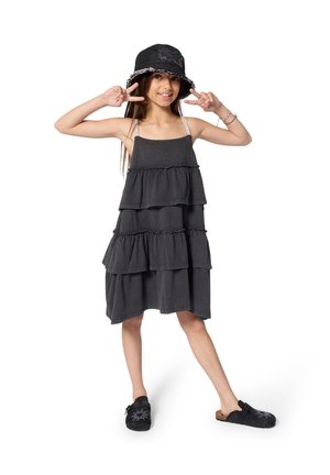 Schwarzes, mehrlagiges Kleid mit Rüschen, dünnen Trägern und lockerem Sitz. Kombiniert mit einem schwarzen Bucket Hat und schwarzen Slippern.