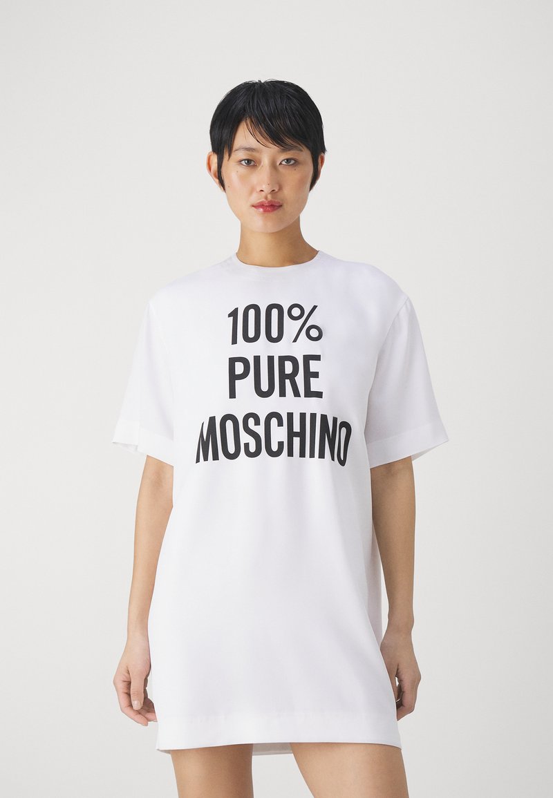 Rochie albă supradimensionată dintr-un material fin, cu mâneci scurte, având un text mare negru care spune "100% PURE MOSCHINO" pe partea din față.
