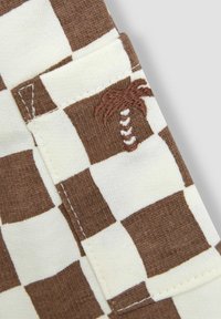 Tela de cuadros marrón y beige con un pequeño detalle bordado de una palmera. Presenta una textura suave y costuras reforzadas en los bordes.